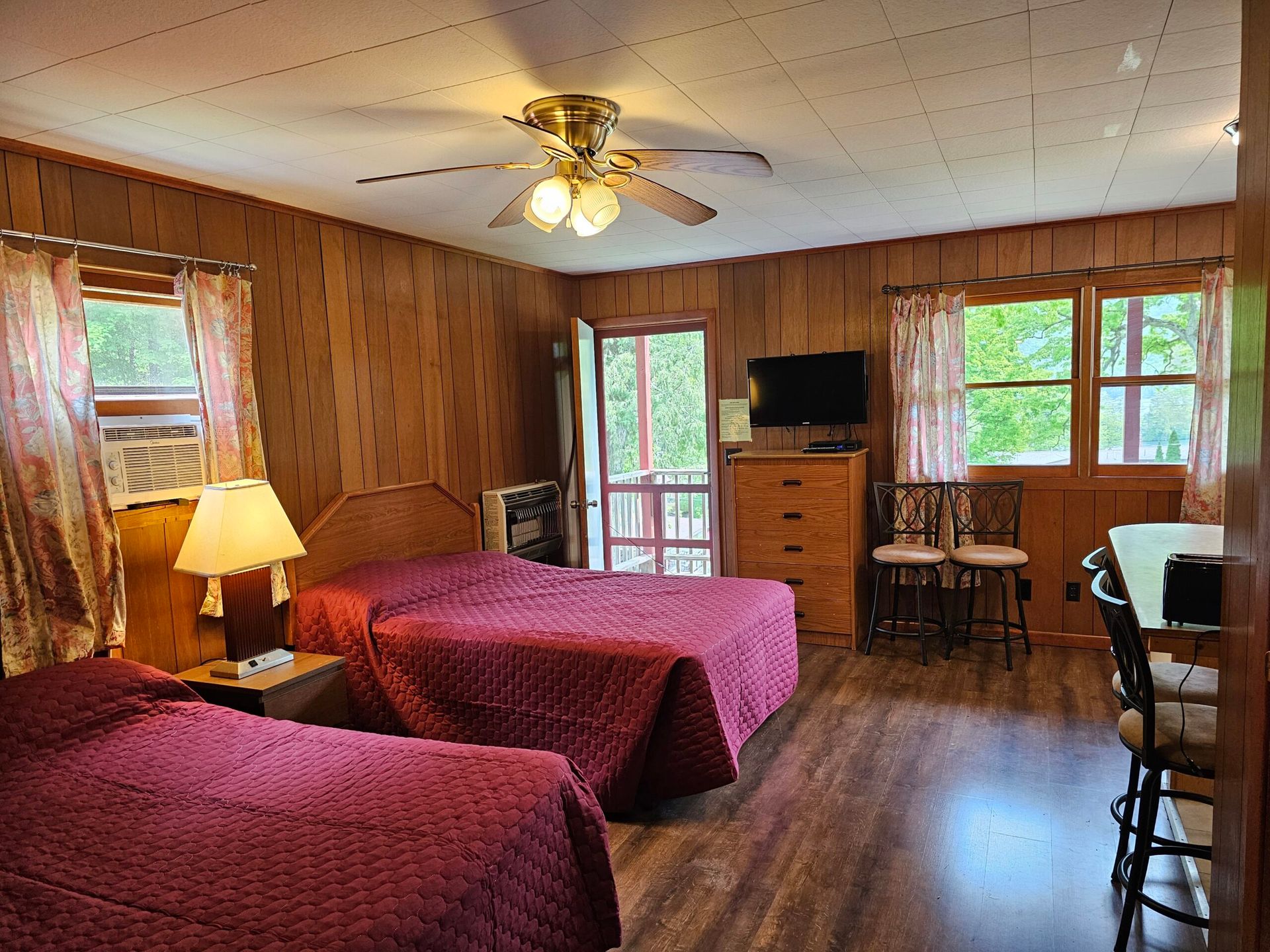 Cozy bedroom featuring two twin beds with maroon bedspreads, wooden paneling, a window with scenic views, and a small kitchen table.